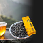 Chén trà phượng hoàng đơn tùng nước vàng óng bên lá trà xoăn dài, nền núi Phượng Hoàng mờ sương